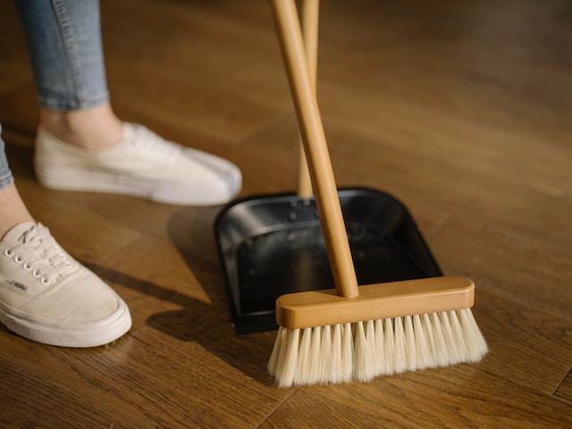 A landlord in white sneakers sweeps the hardwood floor of their rental property with a bamboo broom